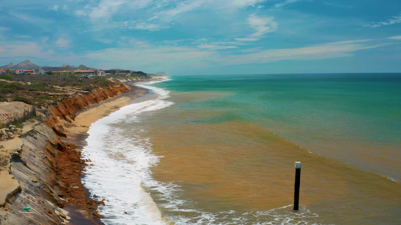 San Jose del Cabo coastline with the surf line angled through the frame, red and tan cliffs to the left, broad turquoise ocean to the right, a black post standing in the water, and no visible people.