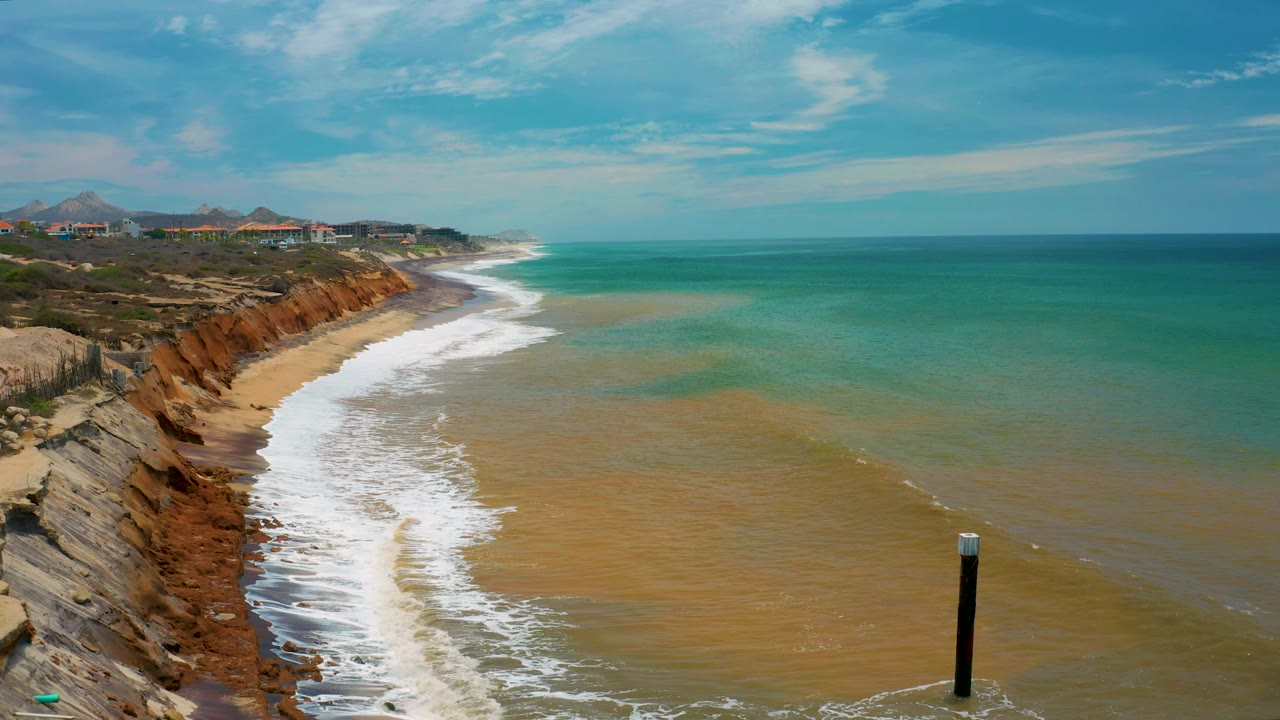 Shoreline below cliffs with surf and a black post near the bottom edge.