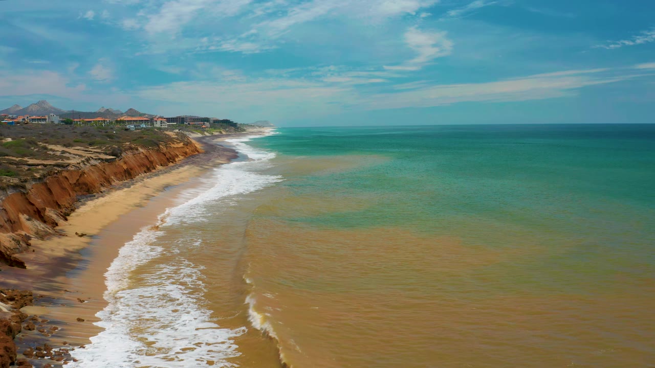 Aerial view of the San Jose Del Cabo beach as a longer white surf band curves along the sand below reddish cliffs, with no visible people.