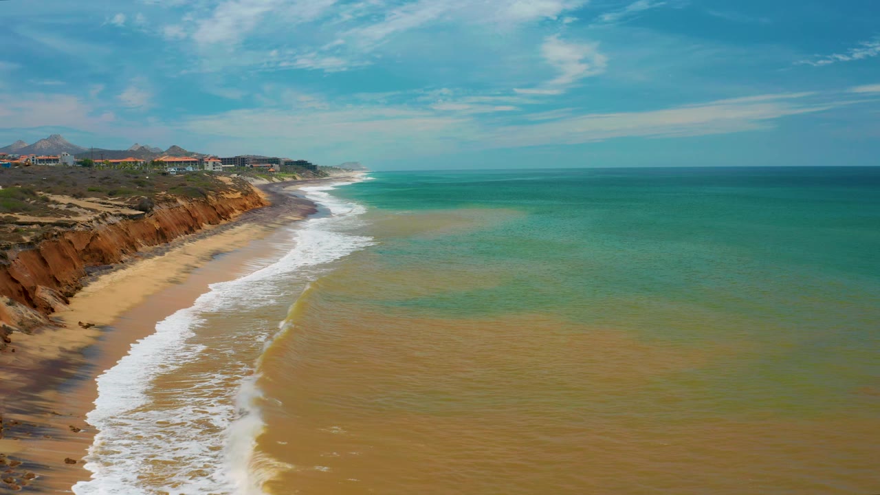 Aerial frame from San Jose Del Cabo showing foamy waves spreading across the lower beach while the ocean opens wide to the right, with no visible people.