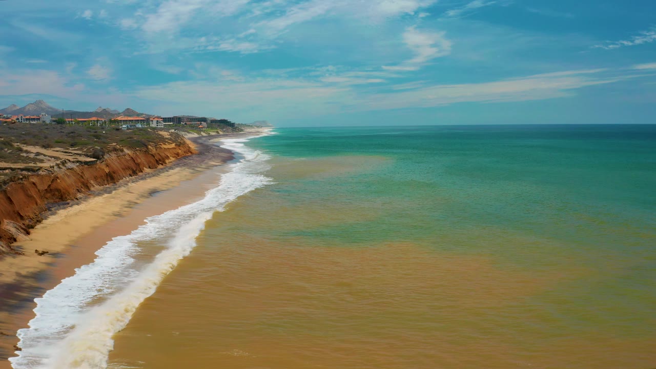 San Jose Del Cabo aerial shoreline frame with the surf band lower in the image, reddish bluffs continuing along the left, and no visible people on the beach.