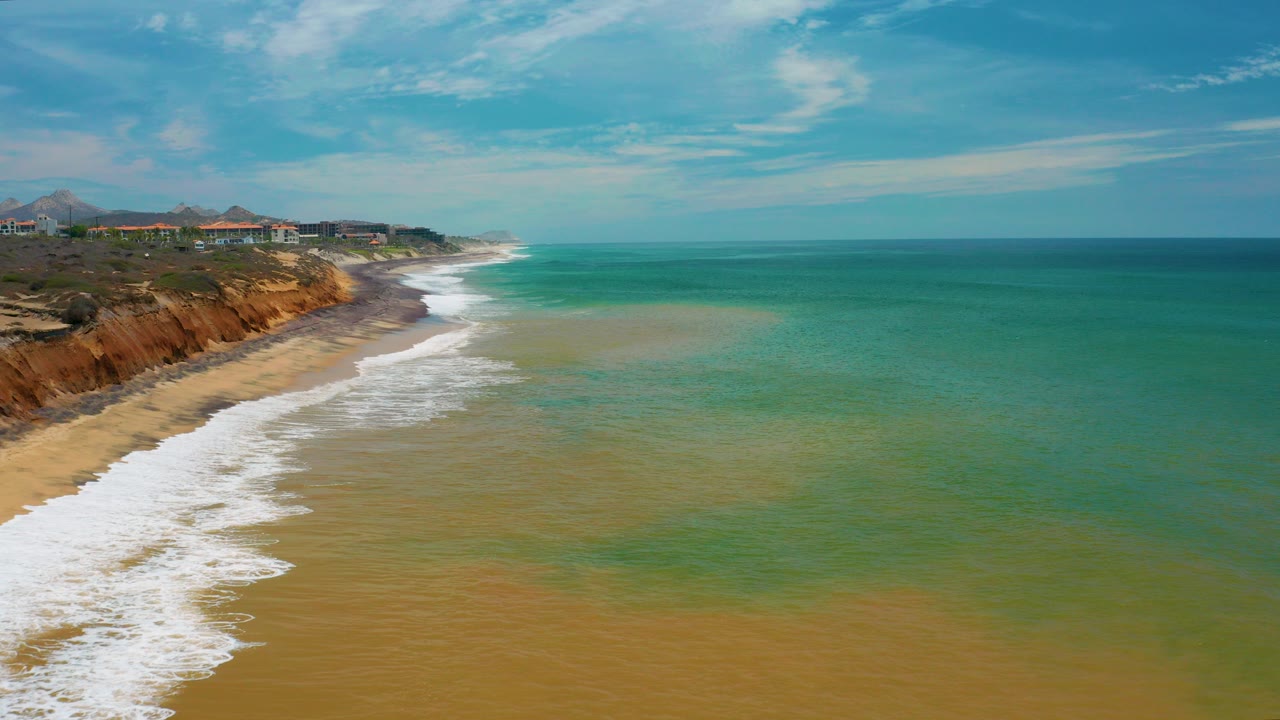 Aerial frame from San Jose Del Cabo, Mexico showing white surf along a sandy beach, reddish cliffs at left, turquoise ocean at right, and no visible people.