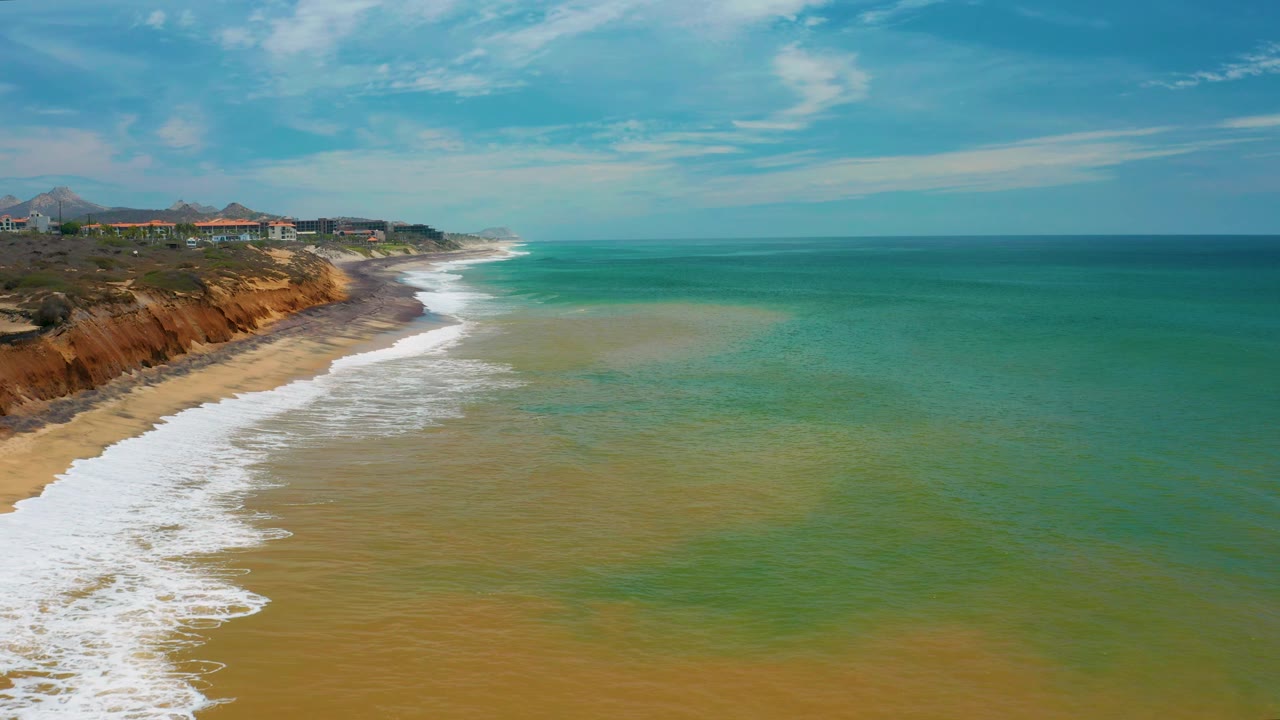 Aerial frame from San Jose Del Cabo, Mexico showing broad bands of tan, green, and blue water beside a sandy beach, low cliffs, distant buildings, and no visible people.