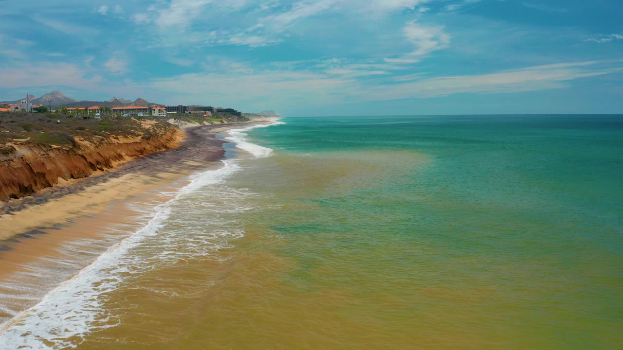 Aerial view in San Jose Del Cabo, Mexico with green sea on the right, white surf reaching a sandy beach, ochre cliffs on the left, distant coastal buildings, hazy hills, and no visible people.