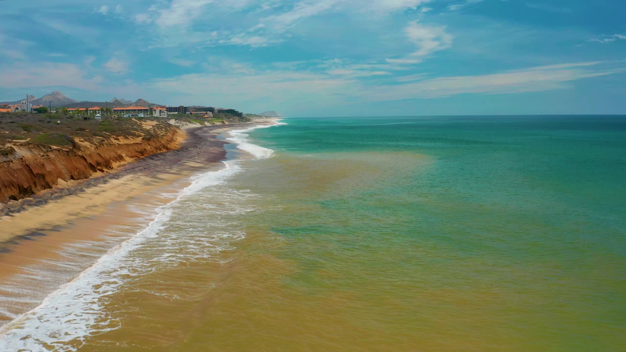 Aerial San Jose Del Cabo coastline with a broad band of green water, foamy waves tracing the beach, orange-brown cliff faces, buildings above the bluff, distant hills, and no visible people.