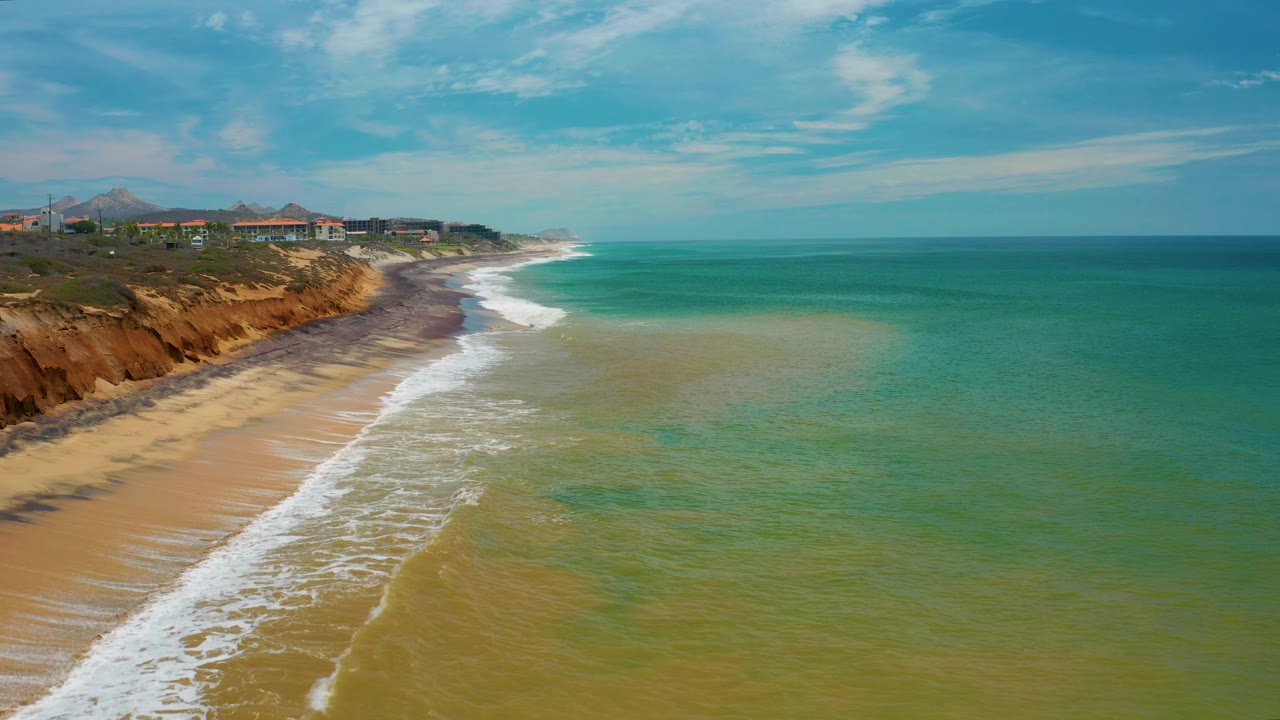 San Jose Del Cabo aerial shoreline with green ocean taking most of the frame, a pale sandy beach below the cliffs, white foam curving toward the distance, low buildings on the bluff, and no visible people.