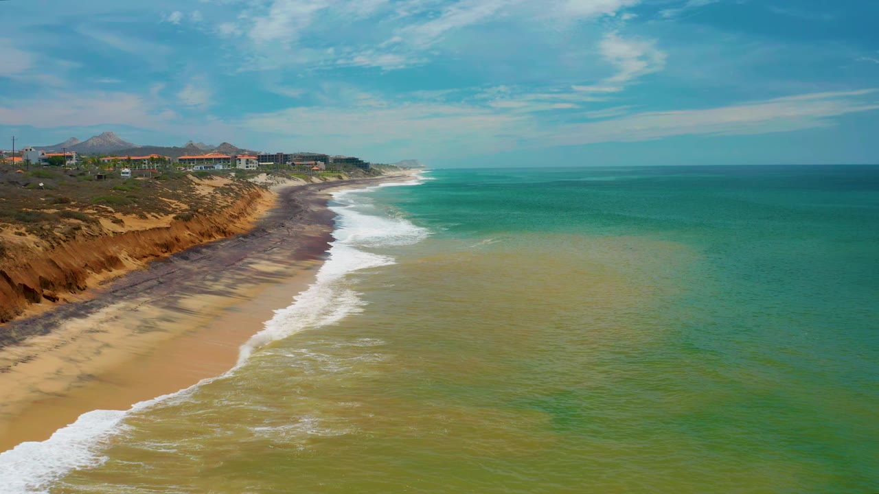 Aerial view along San Jose Del Cabo shoreline, where a wider band of white water reaches the sand below the bluff and the open turquoise ocean fills most of the right side, with no visible people.