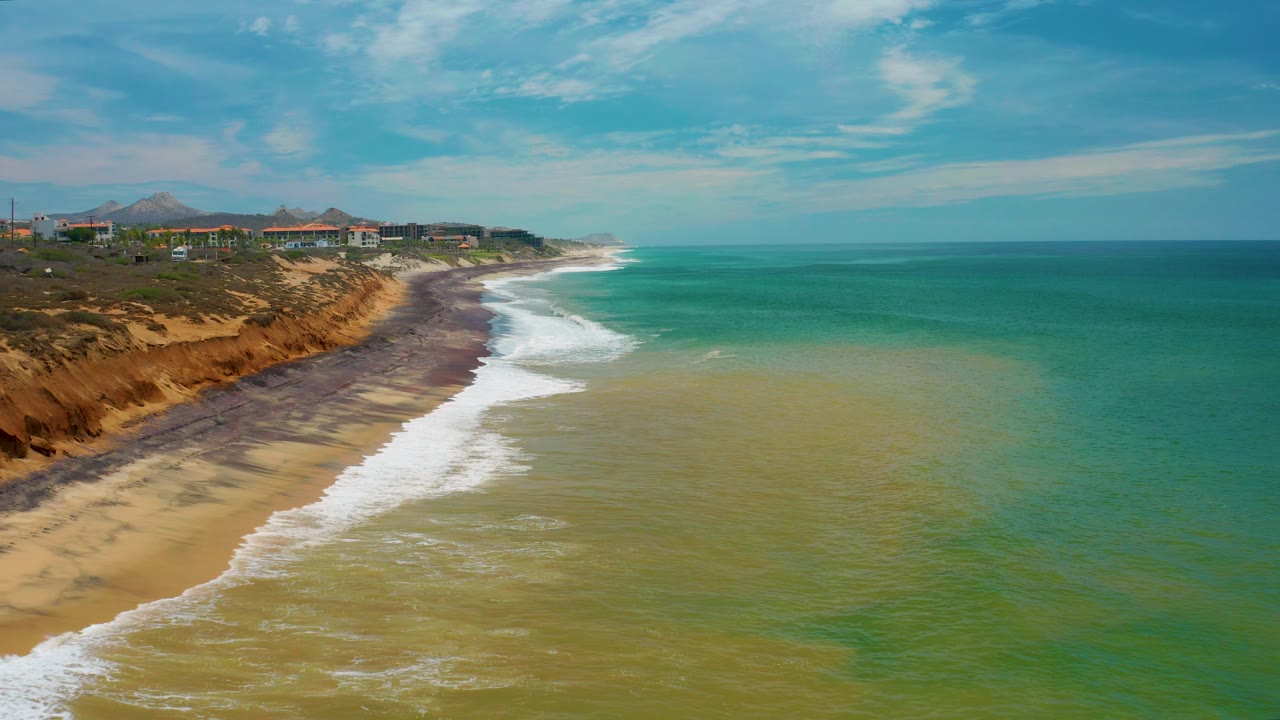 San Jose Del Cabo aerial frame with surf wrapping around the beach edge, darker wet sand near the bluff, green-blue water offshore, ridge-top buildings in the distance, and no visible people.