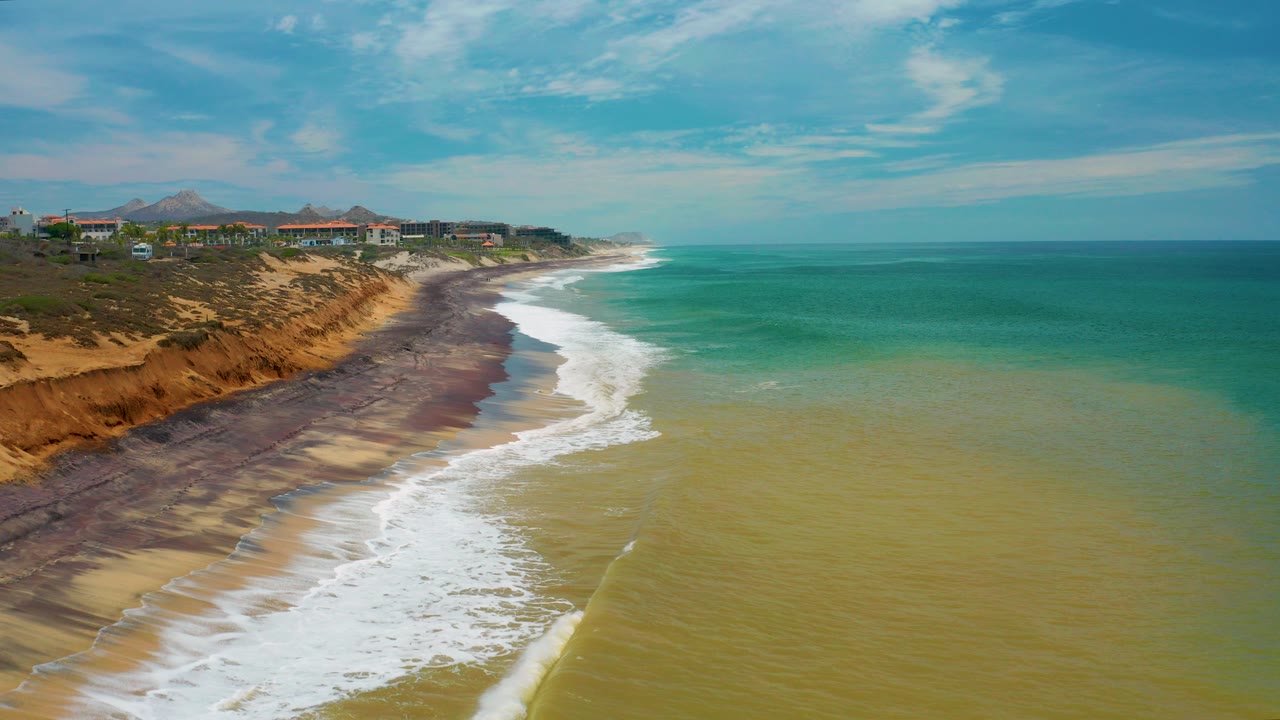 Opening aerial view of the San Jose Del Cabo coastline with white surf curving along tan sand, orange bluffs and scattered buildings on the left, turquoise water on the right, and no visible people.