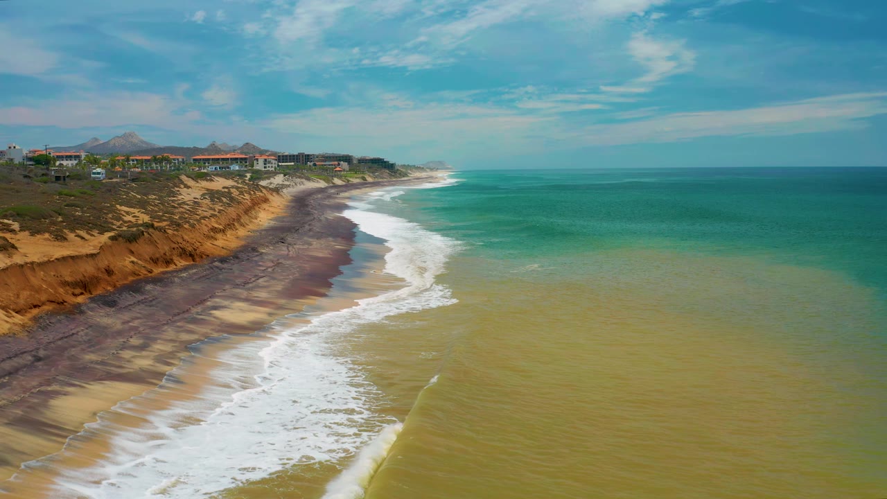 Aerial San Jose Del Cabo beach frame with the foam line slightly farther down the sand, cliffside buildings along the upper left, green-blue water stretching to the horizon, and no visible people.