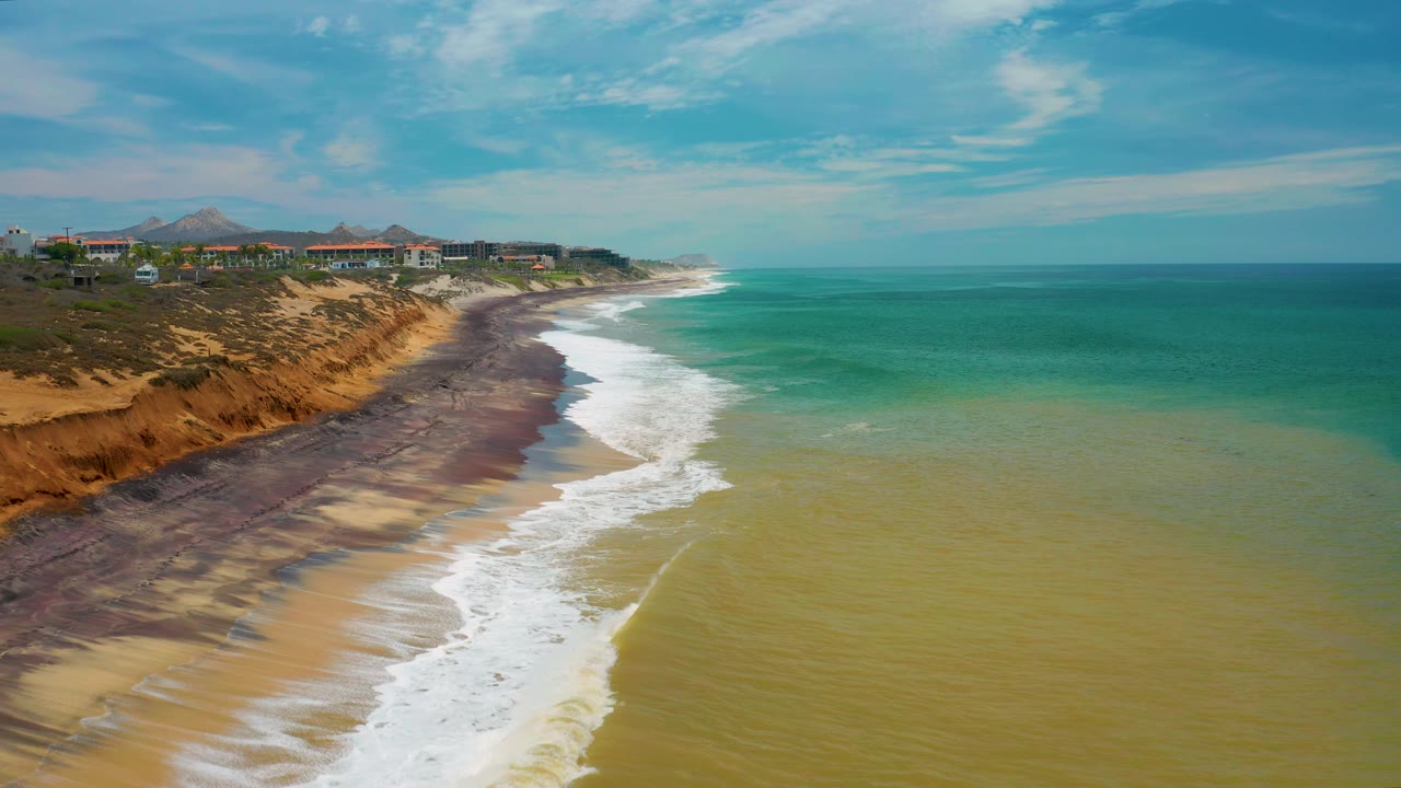 San Jose Del Cabo aerial shoreline view with white water spreading across the lower beach, darker wet sand near the bluff, scattered structures above the coast, open sea to the right, and no visible people.