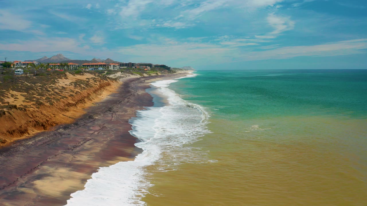 Aerial view in San Jose Del Cabo, Mexico, showing white surf curving along a dark sandy beach below tan cliffs, with coastal buildings and distant hills under clear sky and no visible people.