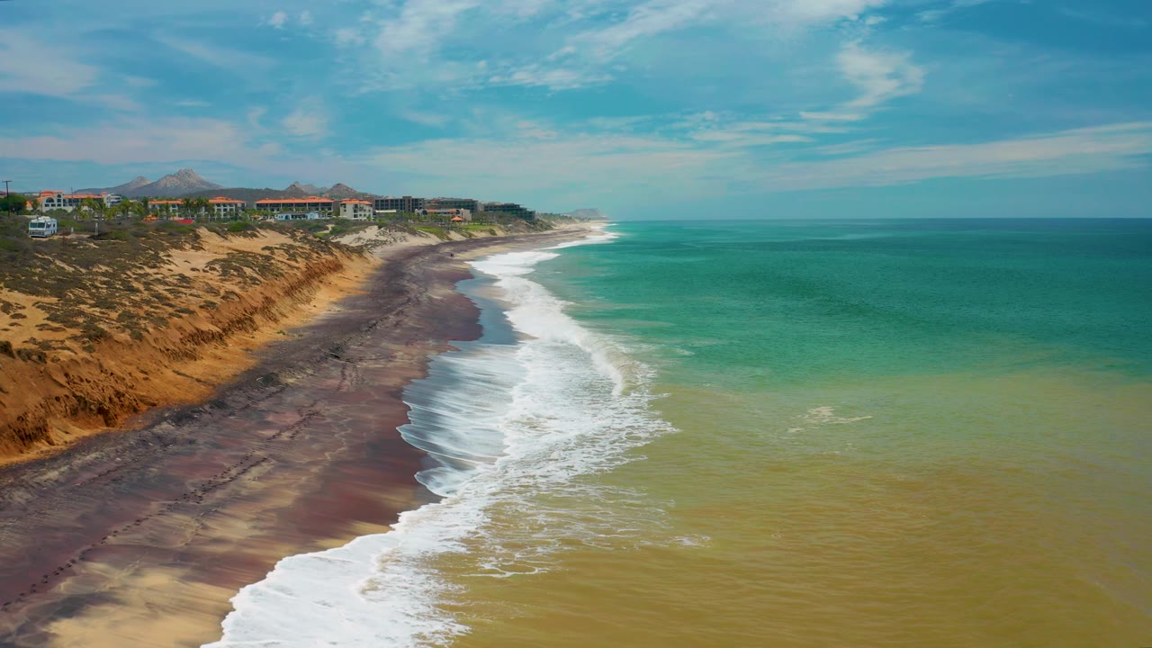 Aerial shoreline frame in San Jose Del Cabo, Mexico, where white breakers roll across dark sand below orange-tan bluffs, with a line of low buildings above the coast and no visible people.