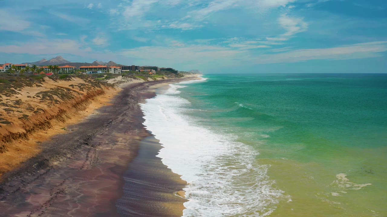 White waves push farther along the dark shoreline in San Jose del Cabo, with tan eroded bluffs and small buildings visible above the beach, and no visible people.