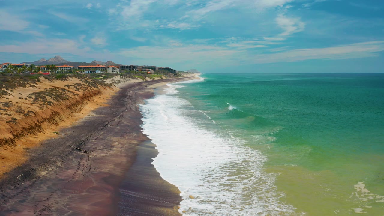 The San Jose del Cabo shore remains framed from above as foamy surf spreads across the beach, green water fills the right side, and no visible people appear on the sand.