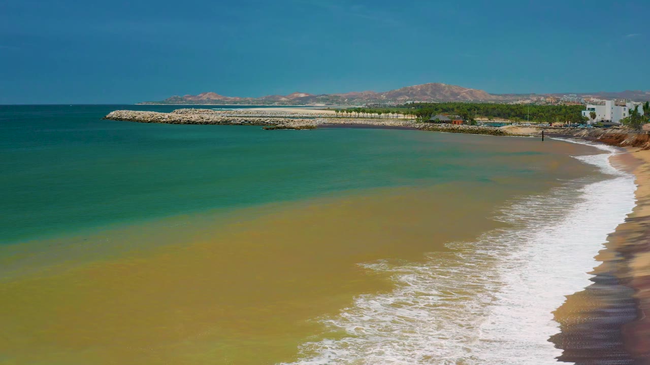 Aerial view of San Jose Del Cabo shoreline with green shallow water, white foam reaching the sand, a rock breakwater, distant hills, and no visible people.