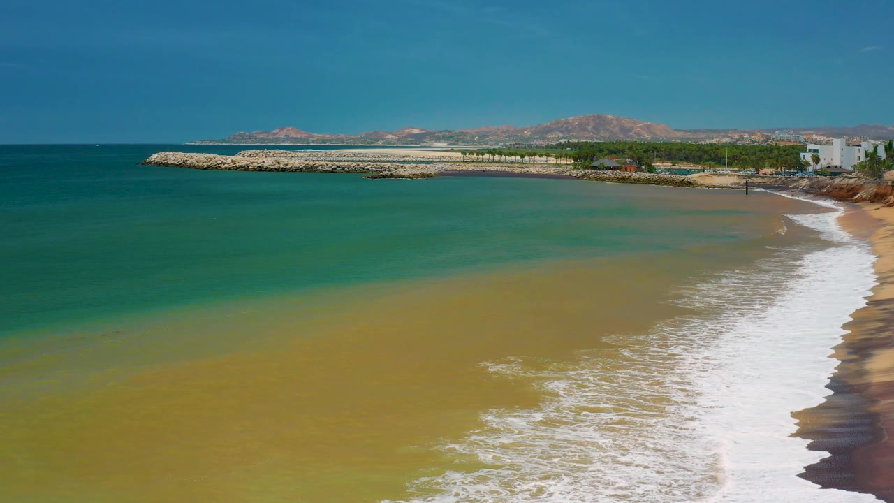 San Jose Del Cabo coastal frame with white surf spreading along the right side of the beach, tan-green water in the foreground, a breakwater in back, and no visible people.