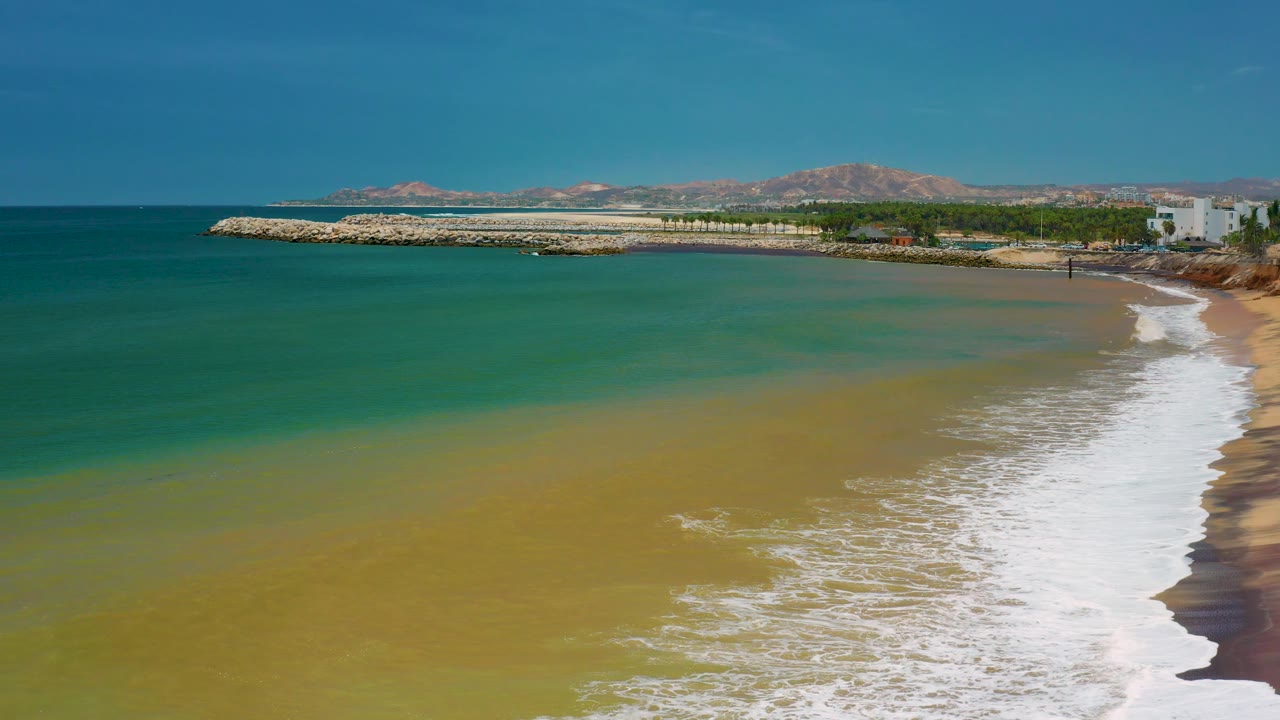 Aerial coastal view with rolling foam, beach sand, blue-green sea, and hills.