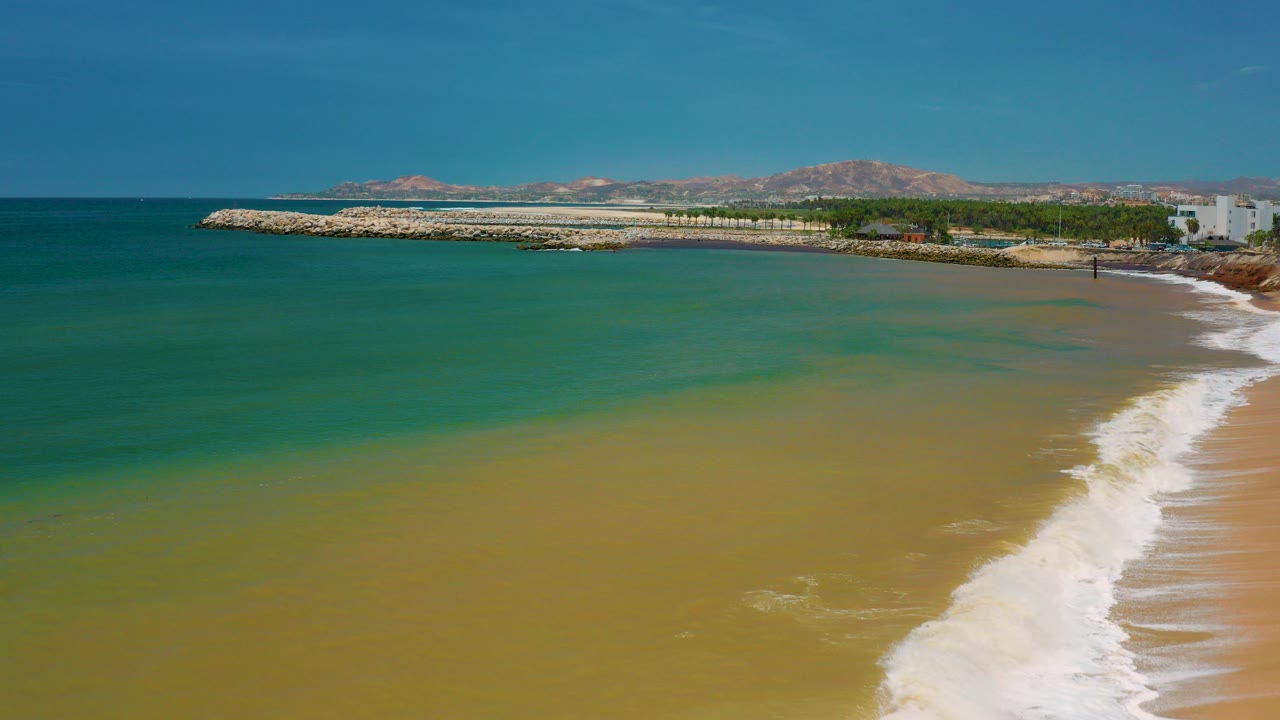 San Jose del Cabo coastline from above with green-blue water in the foreground, a foamy surf line at right, a stone breakwater, distant mountains, and no visible people.