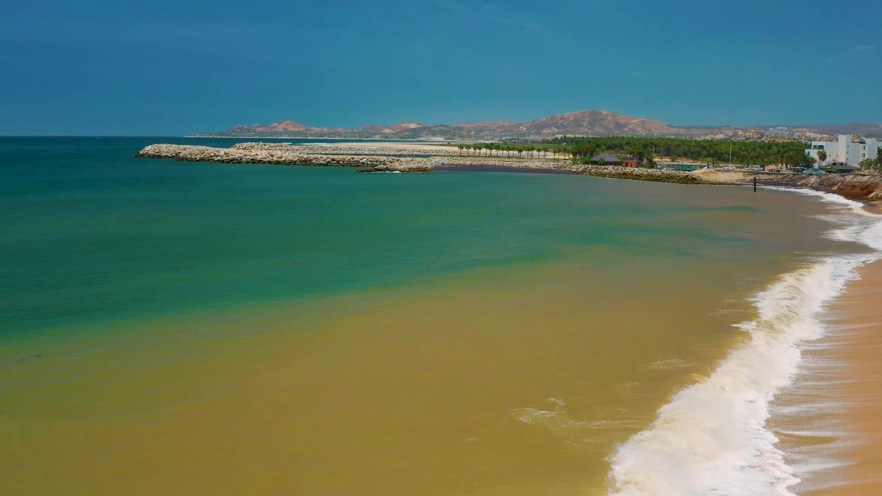 San Jose del Cabo shoreline with teal ocean spread across the frame, pale sandy water near the beach, the breakwater stretching left to right, and no visible people.