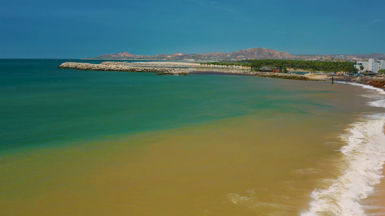 San Jose del Cabo coastal frame with more sandy-colored shallows in the lower right, green ocean to the left, the breakwater across the midground, mountains beyond, and no visible people.
