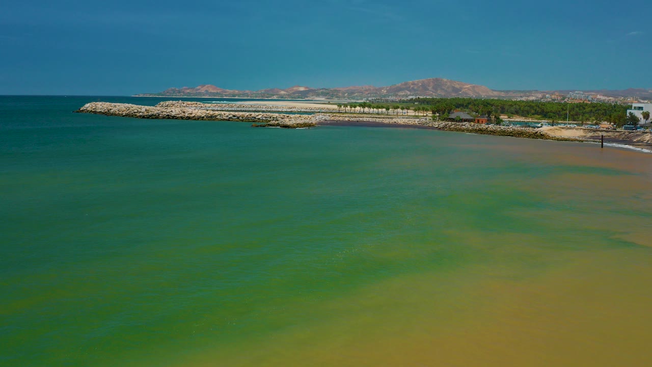 Aerial view over green ocean water toward the San Jose del Cabo coastline, with a rocky breakwater, pale beach shallows, distant mountains, blue sky, and no visible people.