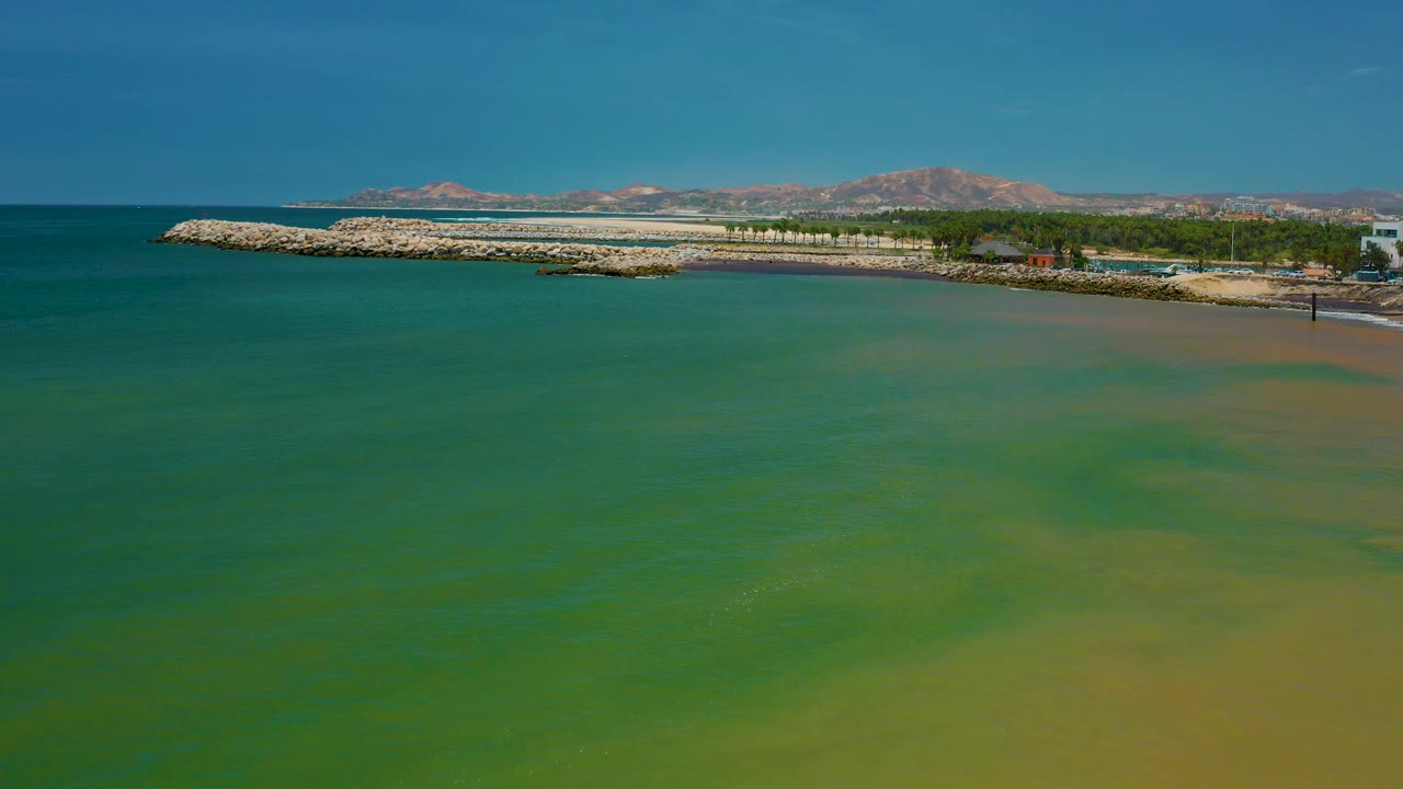 Aerial view of green Pacific water approaching a curved rock barrier on the San Jose del Cabo coast, with sandy shallows, low buildings or structures at the shore, mountains, and no visible people.