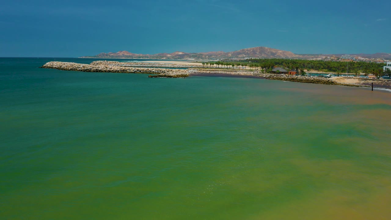Aerial frame in San Jose del Cabo with broad green water in the foreground, a rocky coast and thin beach line in the upper frame, distant mountains, clear blue sky, and no visible people.
