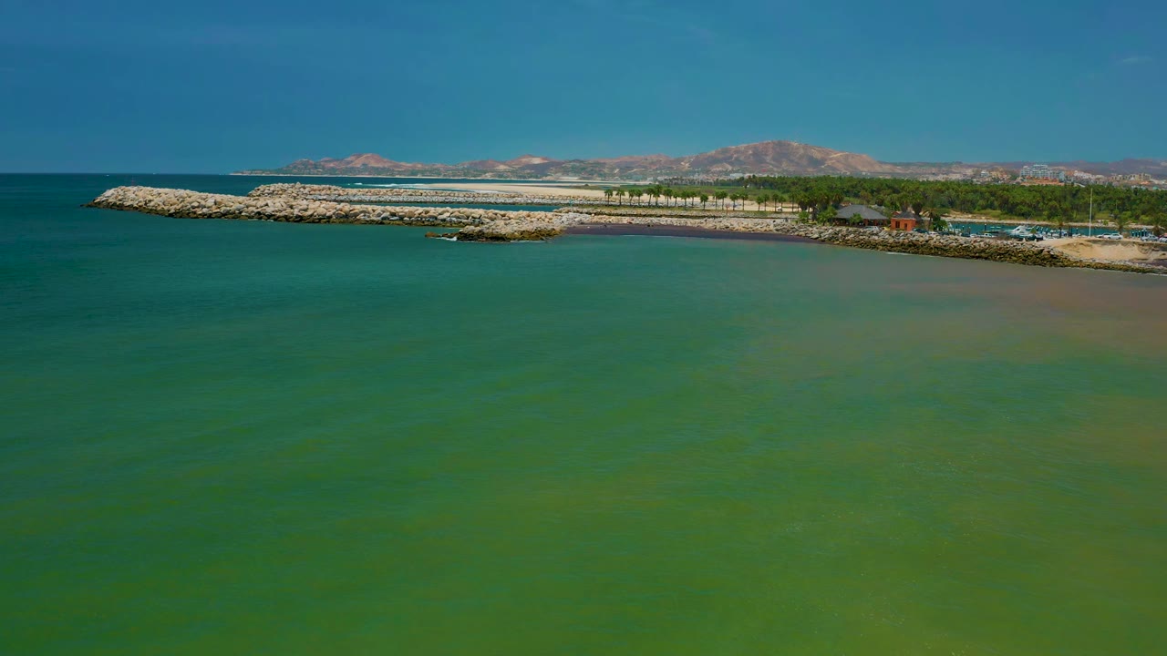 San Jose Del Cabo shoreline seen from above, with green water taking most of the frame, a rocky barrier near the horizon, trees and small structures behind it, and no visible people.