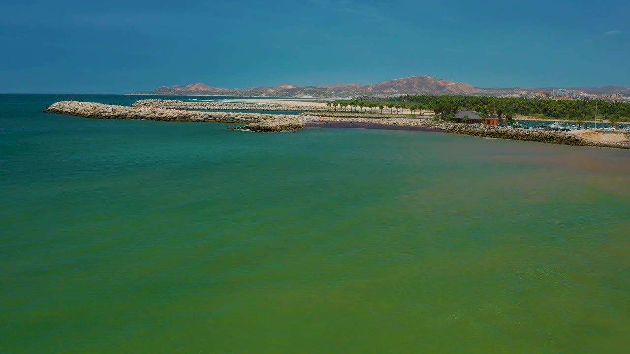 A broad aerial frame of San Jose Del Cabo green seawater, with a rocky seawall and marina-side shoreline running across the background, low trees, distant mountains, and no visible people.