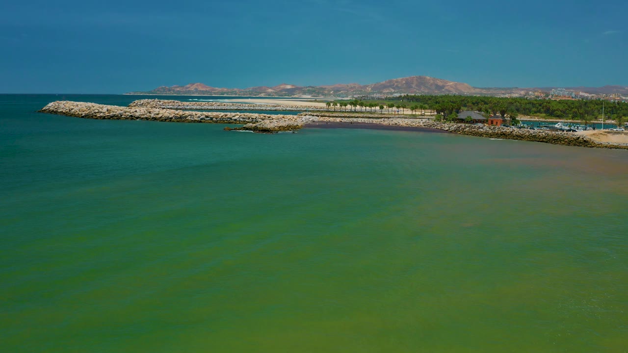 Green seawater stretches toward a pale rock seawall in San Jose Del Cabo, with a tree-lined shore, low buildings, faint parked vehicles or boats, brown mountains, and no visible people.