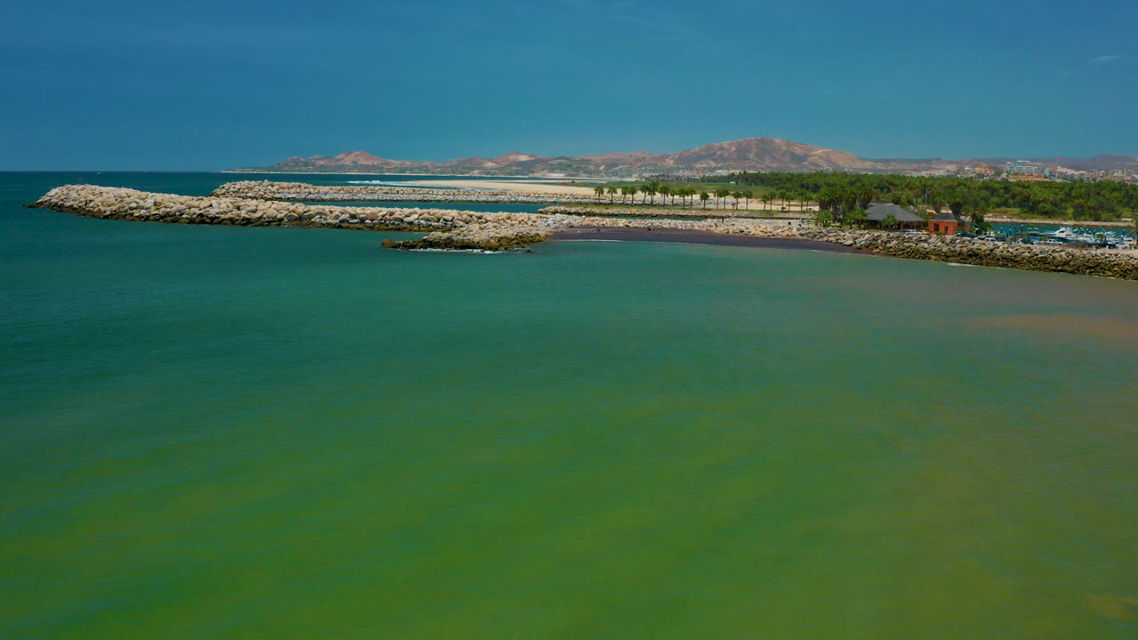 Wide aerial frame from San Jose Del Cabo, Mexico, looking over green turquoise water toward pale rock breakwaters, a small rocky islet, shoreline trees, distant low buildings, mountains, and no visible people.