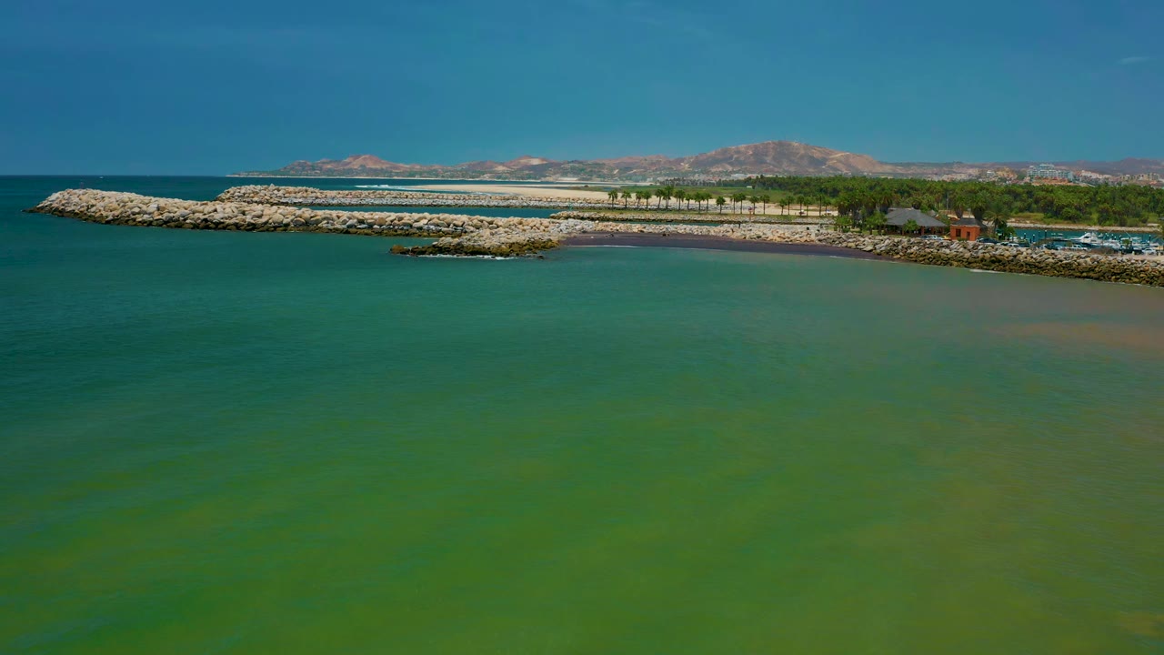 Aerial coastline view with turquoise water, rock barriers, trees, and mountains in the distance.