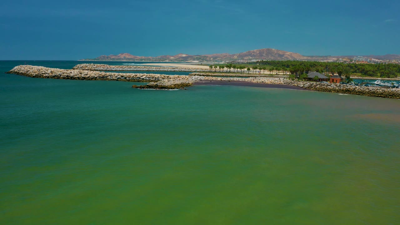 Drone frame over San Jose Del Cabo water with the rock breakwater running across the coast, a small rocky outcrop near shore, trees and low development on the right, mountains on the horizon, and no visible people.