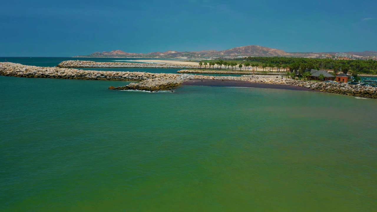 Second frame holds a wide aerial view over San Jose del Cabo water, with a rocky outcrop near the center, a longer breakwater behind it, trees to the right, and no visible people.