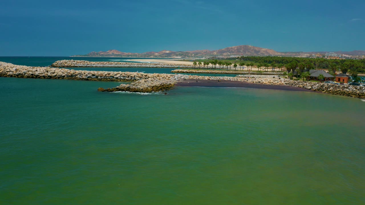 Mid-clip frame in San Jose del Cabo shows a broad foreground of teal water, a rocky spit left of center, a sandy shoreline, trees and low structures at right, and no visible people.