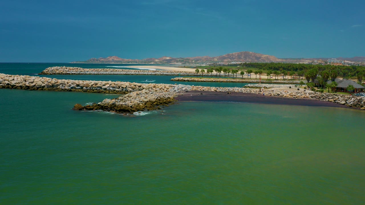 Opening aerial frame over San Jose Del Cabo, Mexico, with green water in front, pale stone breakwaters across the shore, a palm line, distant dry hills, and no visible people.