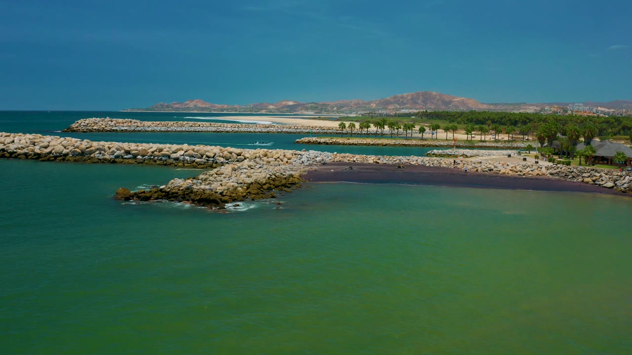 Aerial view in San Jose Del Cabo showing green sea in the foreground, a broad line of white rocks, a darker beach strip, palm trees to the right, distant hills, and no visible people.