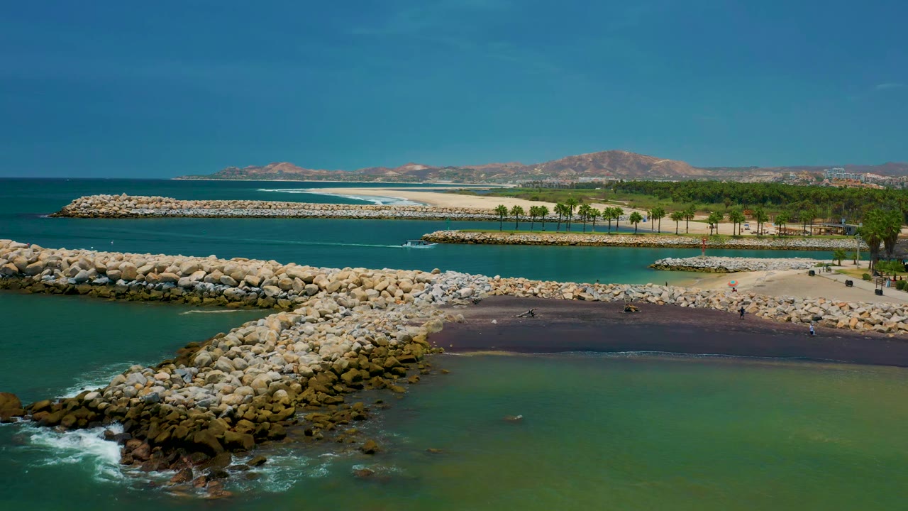 Rock jetty and sheltered coastal water with palms, sand, surf, and mountains beyond.