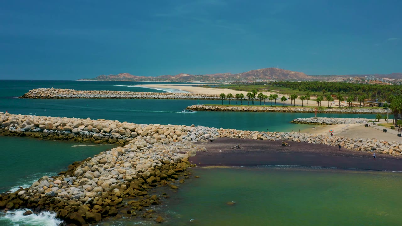 Aerial view in San Jose del Cabo with breakwater rocks crossing the foreground, calm green inlet water, a narrow sandy beach with palms, white surf at the outer shore, and low mountains across the horizon.