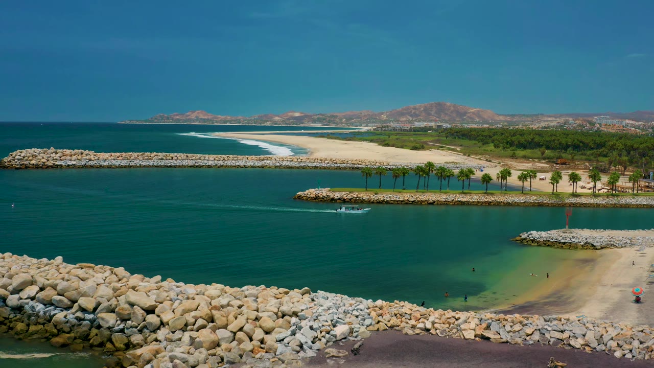 San Jose del Cabo aerial frame with the small white boat farther along the green-blue channel, rock jetties in the foreground, sandy beach beyond, and no clearly visible people.