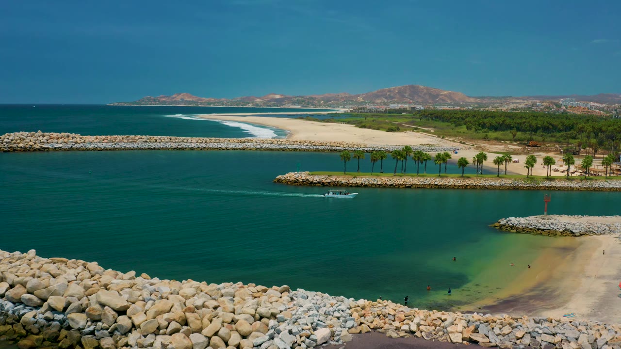 Elevated San Jose del Cabo view with the boat centered below palm-lined sand, a white surf line on the outer beach, rock breakwaters, and no clearly visible people.