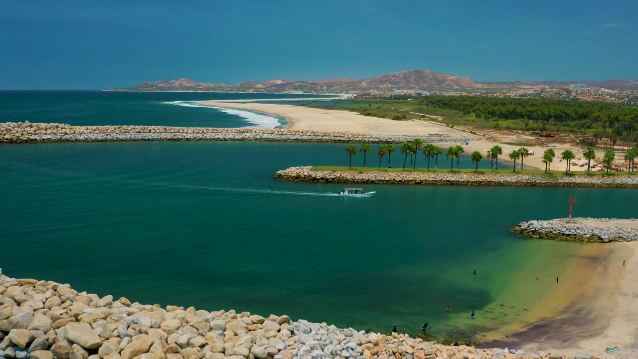 Wide San Jose del Cabo coastal aerial showing the boat moving across the channel below a row of palms, with foreground rocks, a sandy beach, distant hills, and no clearly visible people.