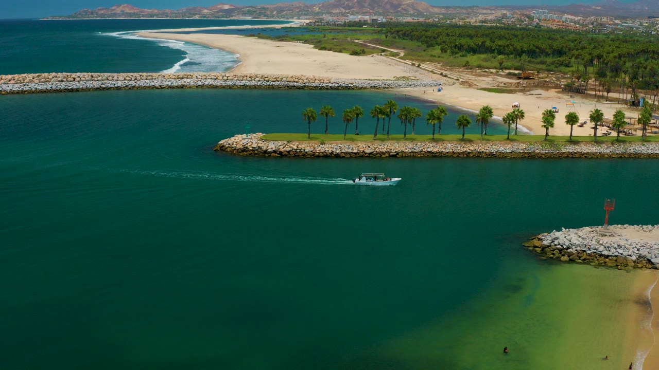 Aerial view of a small boat crossing teal water near a beach and rock jetties.