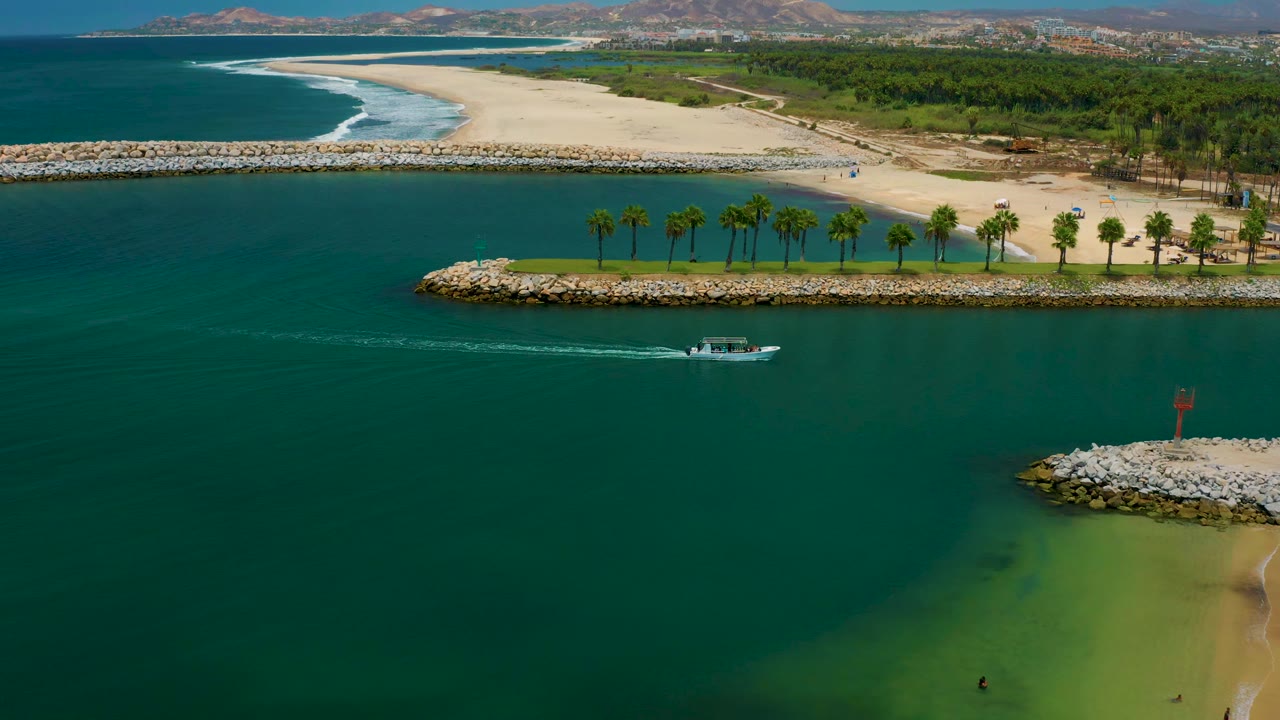Aerial frame from San Jose Del Cabo, Mexico with the small boat slightly farther across blue-green water, a thin wake behind it, rock barriers, palms, beach sand, surf, and no visible people.