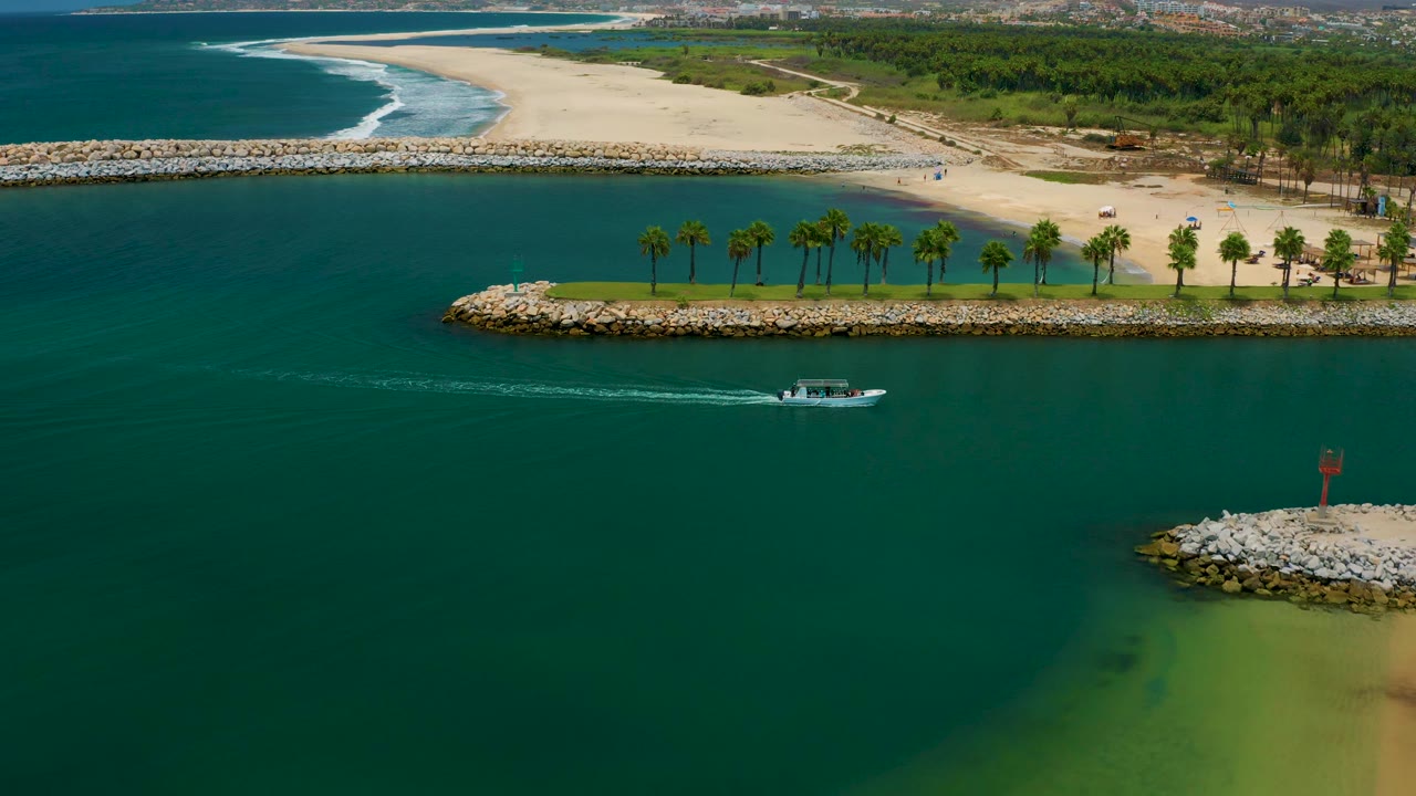 Aerial frame from San Jose Del Cabo, Mexico showing the boat farther right on open teal water, with white surf curving along the beach, a palm strip on rocks, and no visible people.