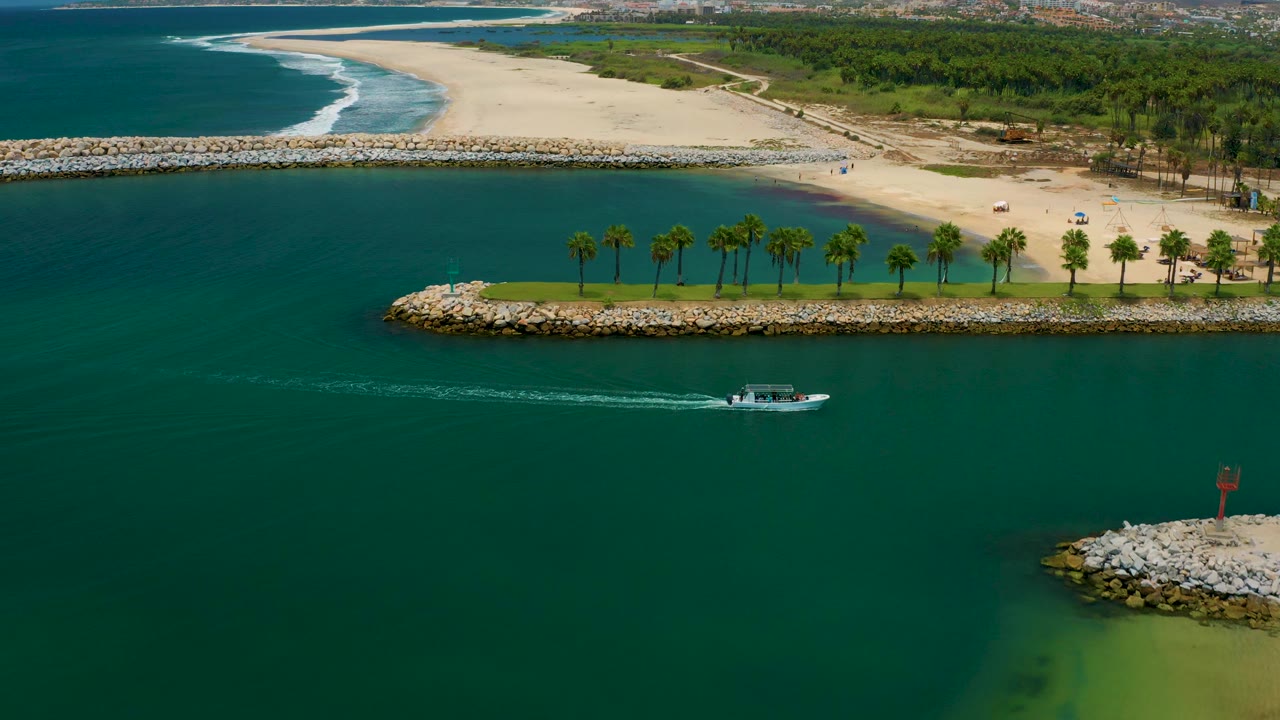Aerial frame from San Jose Del Cabo, Mexico with the boat low in frame, a long wake across dark teal water, palm trees on a narrow green breakwater, sandy beach, surf, and no visible people.