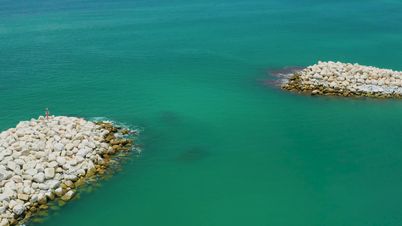 Aerial frame from San Jose del Cabo, Mexico, with the nearer rock breakwater shifted farther into view at lower left, a small marker on the stones, and no visible people.