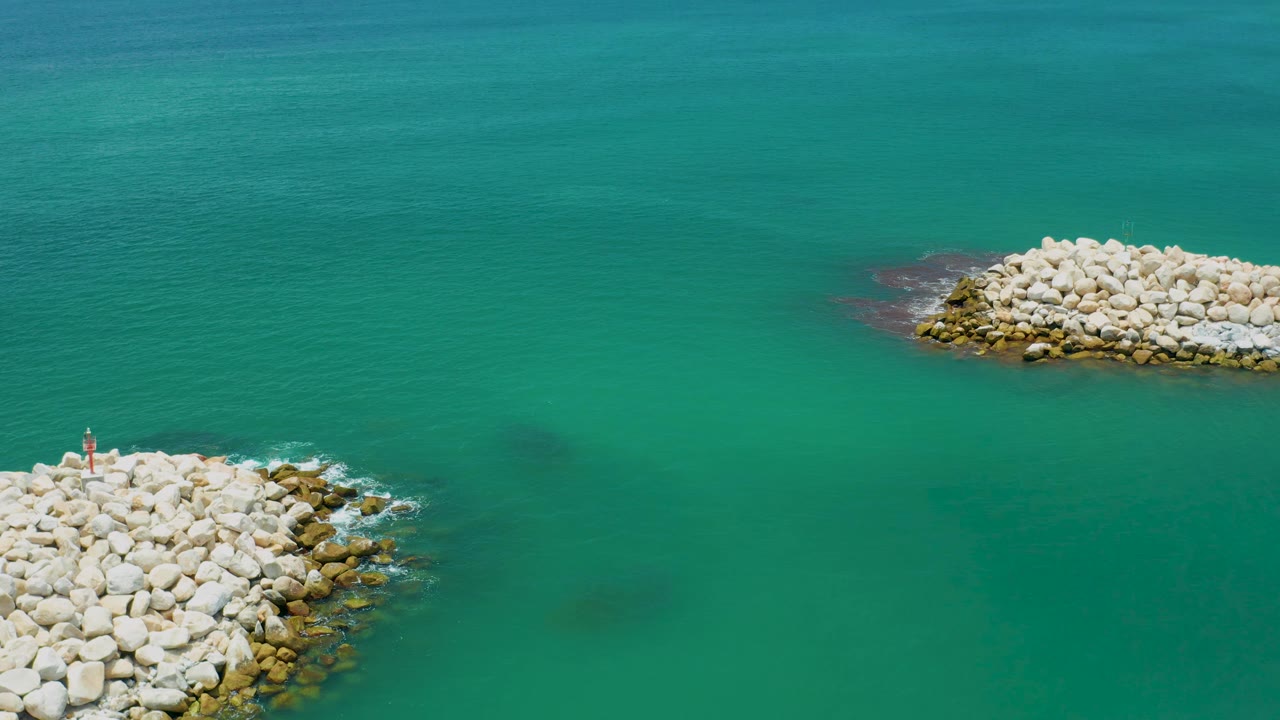 Aerial frame in San Jose Del Cabo, Mexico, showing blue-green water, a lower-left stone jetty with a small red-and-white marker, and a pale rock pile near the upper center, with no visible people.