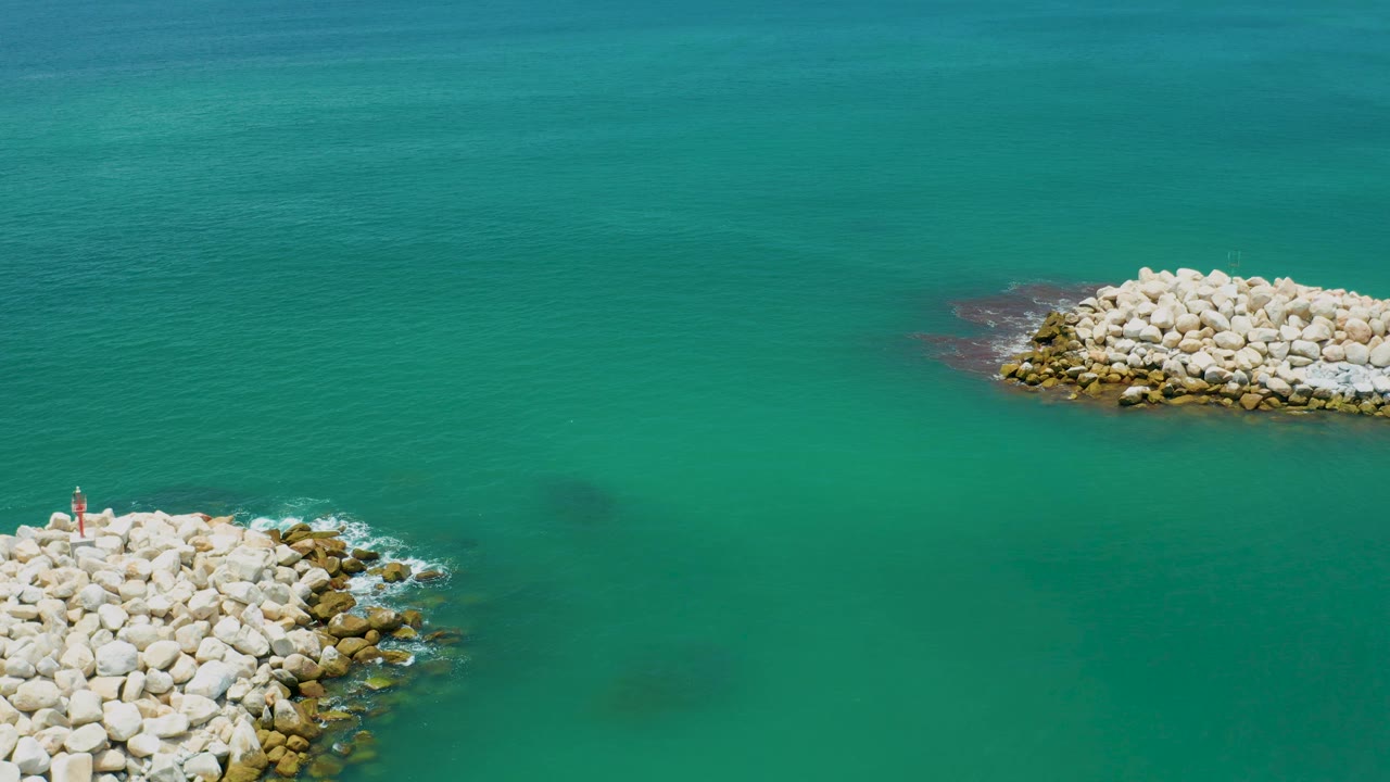 Aerial view in San Jose Del Cabo, Mexico, with clear teal water between two pale rock breakwaters and faint dark areas below the surface, with no visible people.